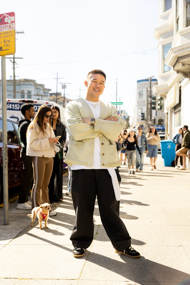 A smiling man stands confidently with arms crossed on a sunny sidewalk, while people and a small dog in a red harness gather behind him.